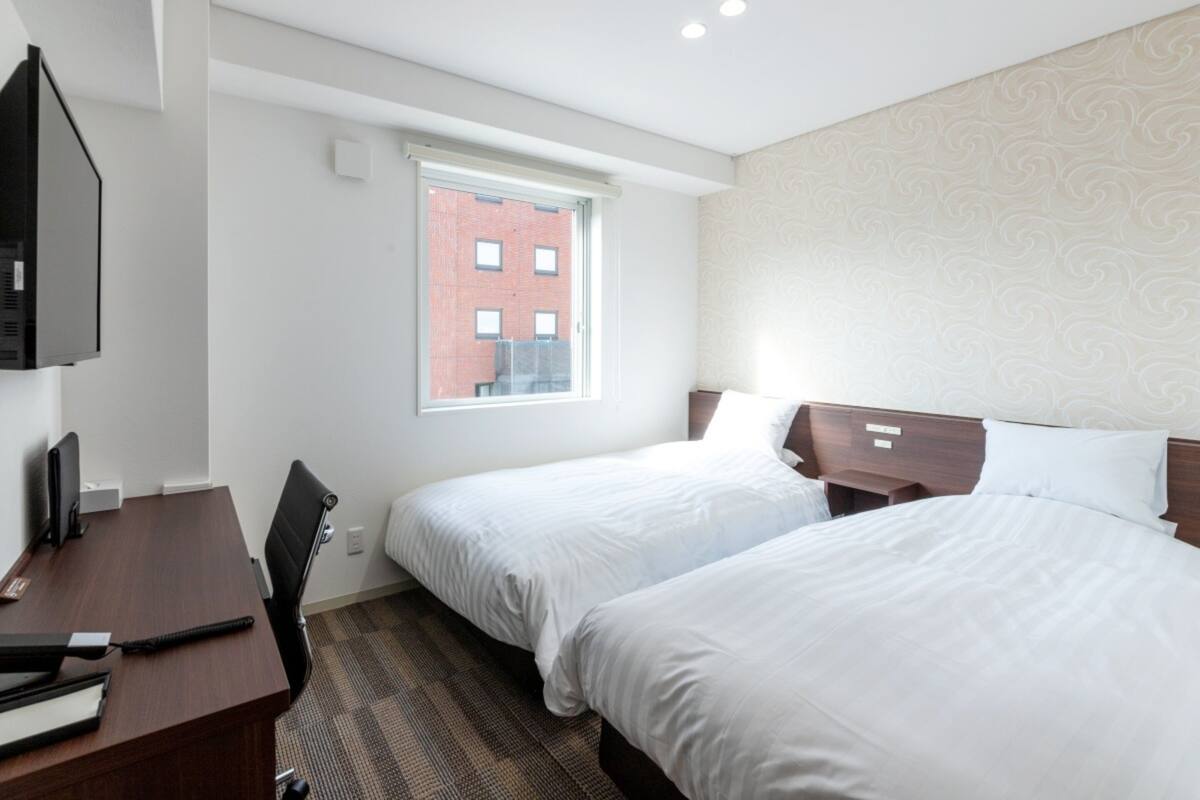 Une chambre d’hôtel avec deux lits, un bureau, une chaise, une télévision et une fenêtre donnant sur un immeuble en briques.