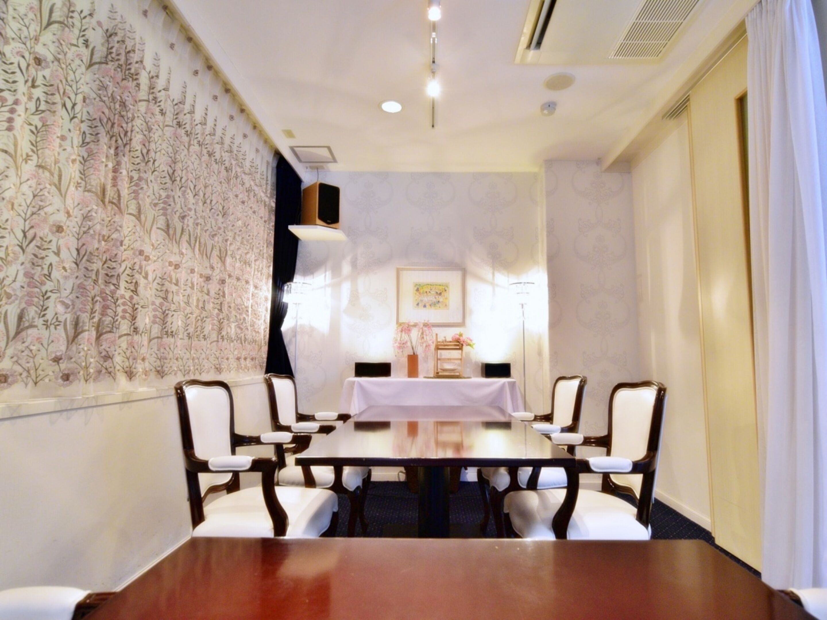 A conference room with a long table, white chairs, floral wallpaper, a framed picture on the wall, and a ceiling light.