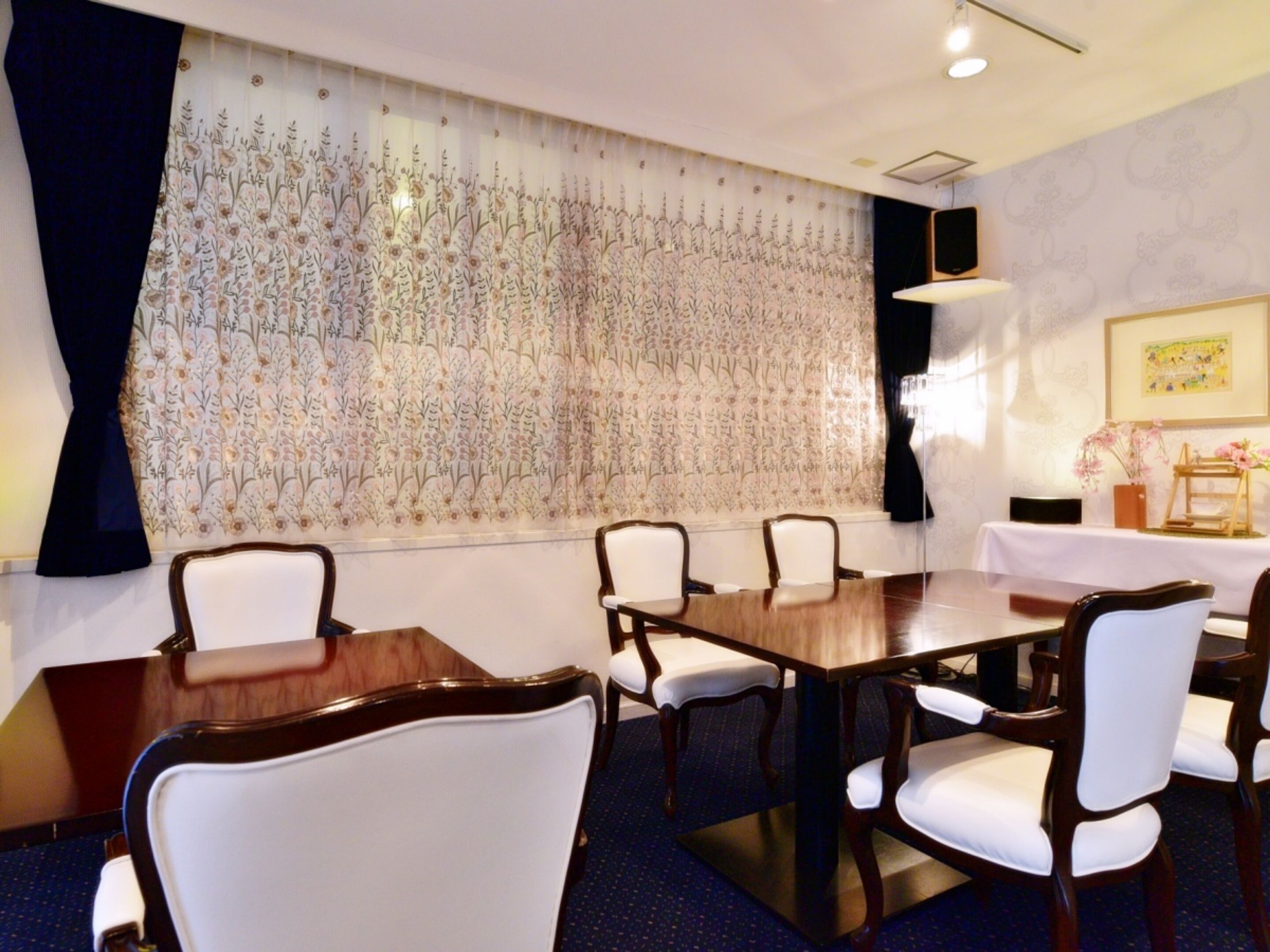 A conference room with a large table, white upholstered chairs, and a floral-patterned wall.