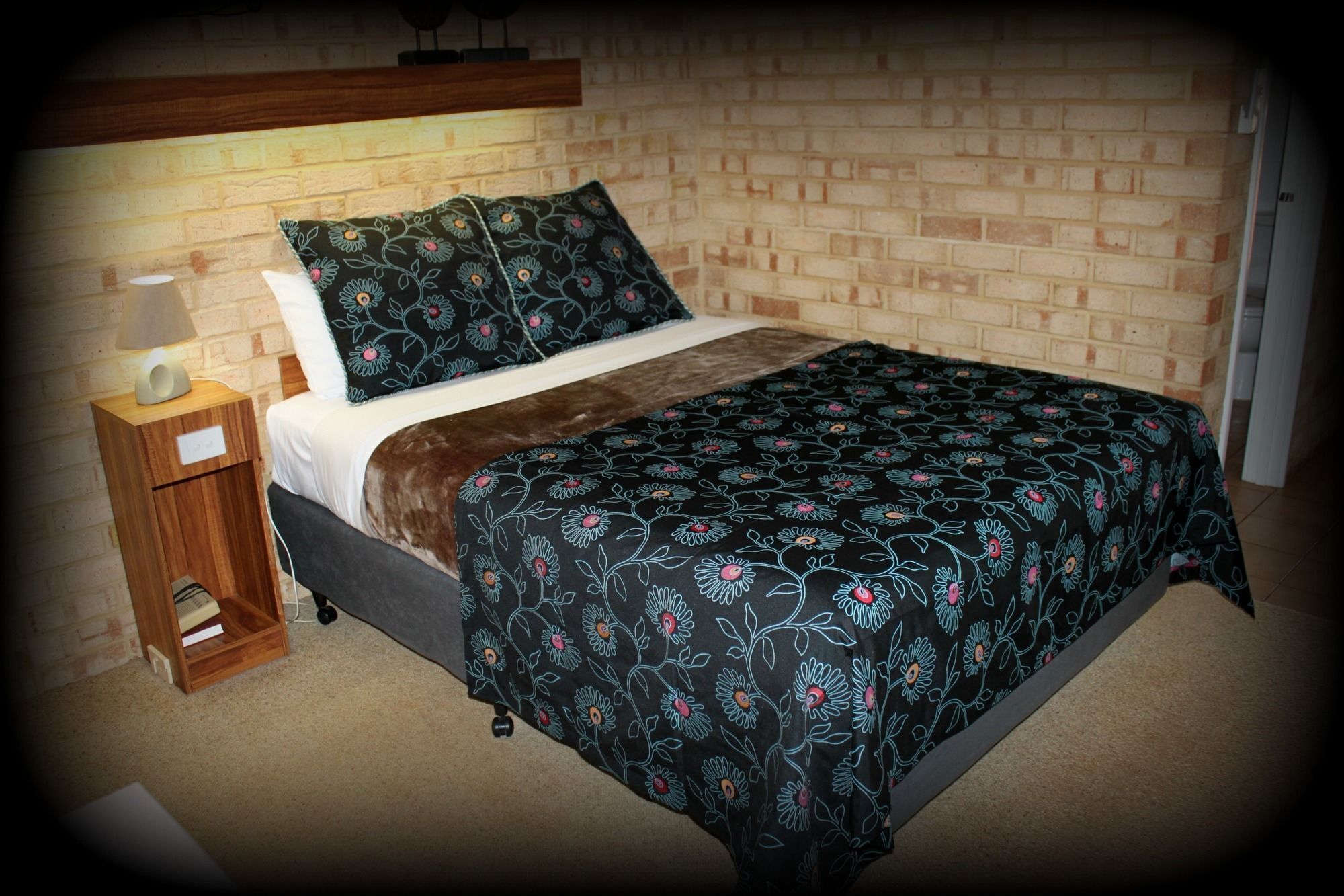 Een slaapkamer met een bakstenen muur, een houten nachtkastje met een lamp, een bed met een bloemenlaken en een hoofdbord.