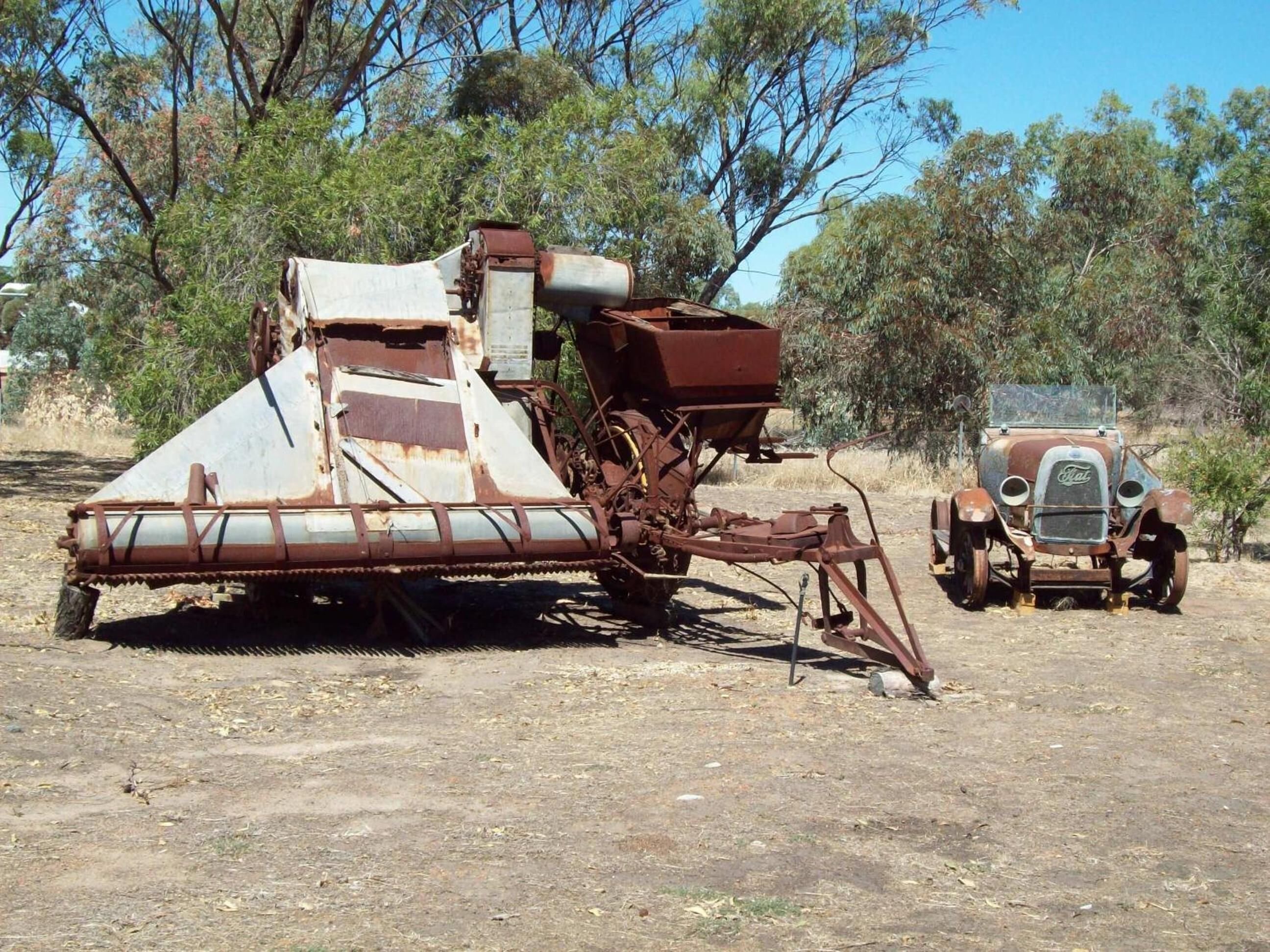 An old, rusted piece of machinery with a trailer, surrounded by dry, sparse vegetation and a clear sky.