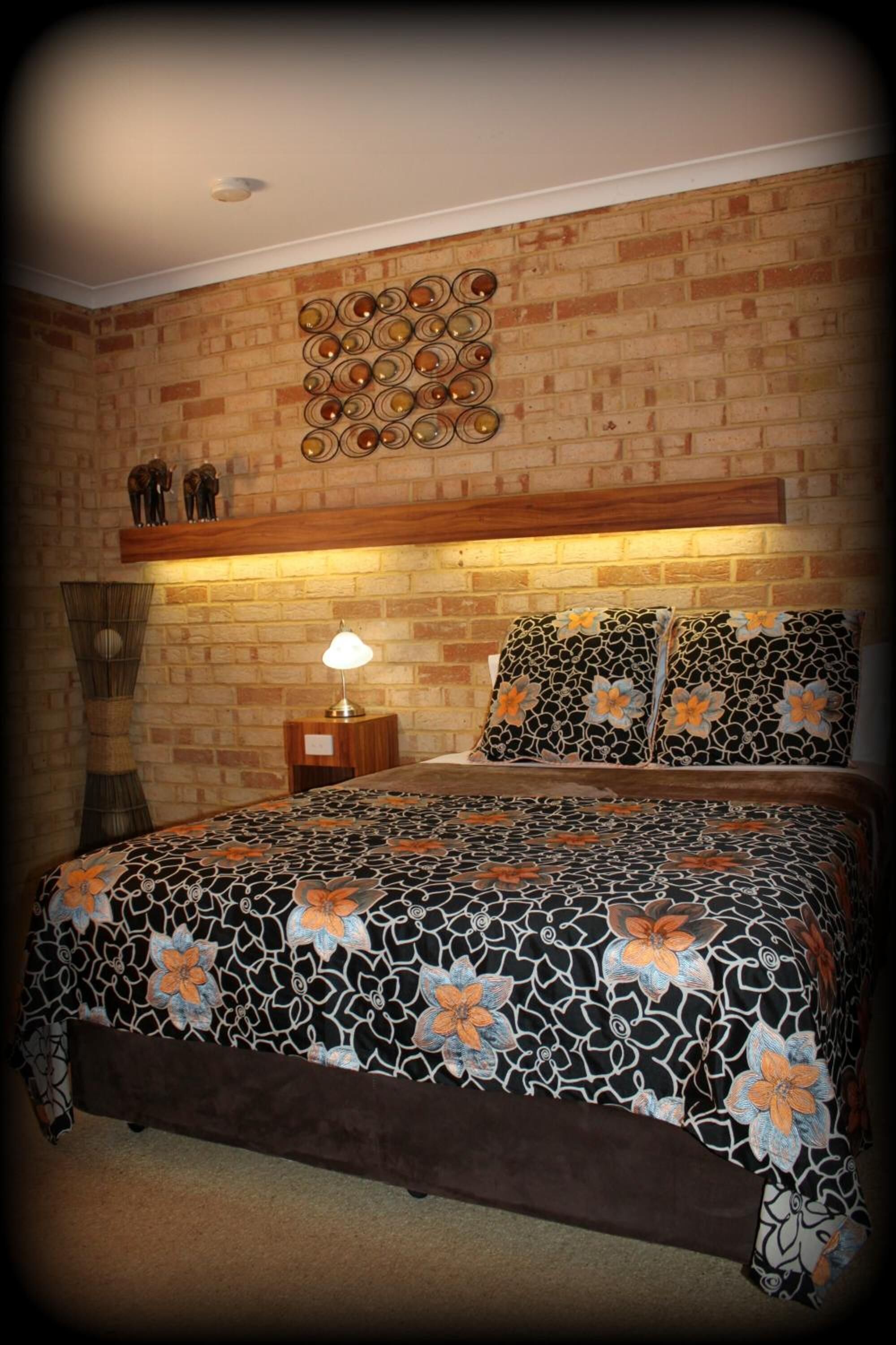 Ein Schlafzimmer mit Ziegelwand, hölzernem Kopfteil, einem Bett mit gemustertem Überwurf, zwei Kissen mit Blumenmuster, einer Nachttischlampe und einem Wanddekor.