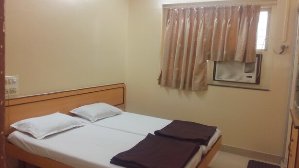 A single bed with white linens and two pillows, a window with curtains, and an air conditioning unit on the wall. - Milan Guest House (Mumbai)