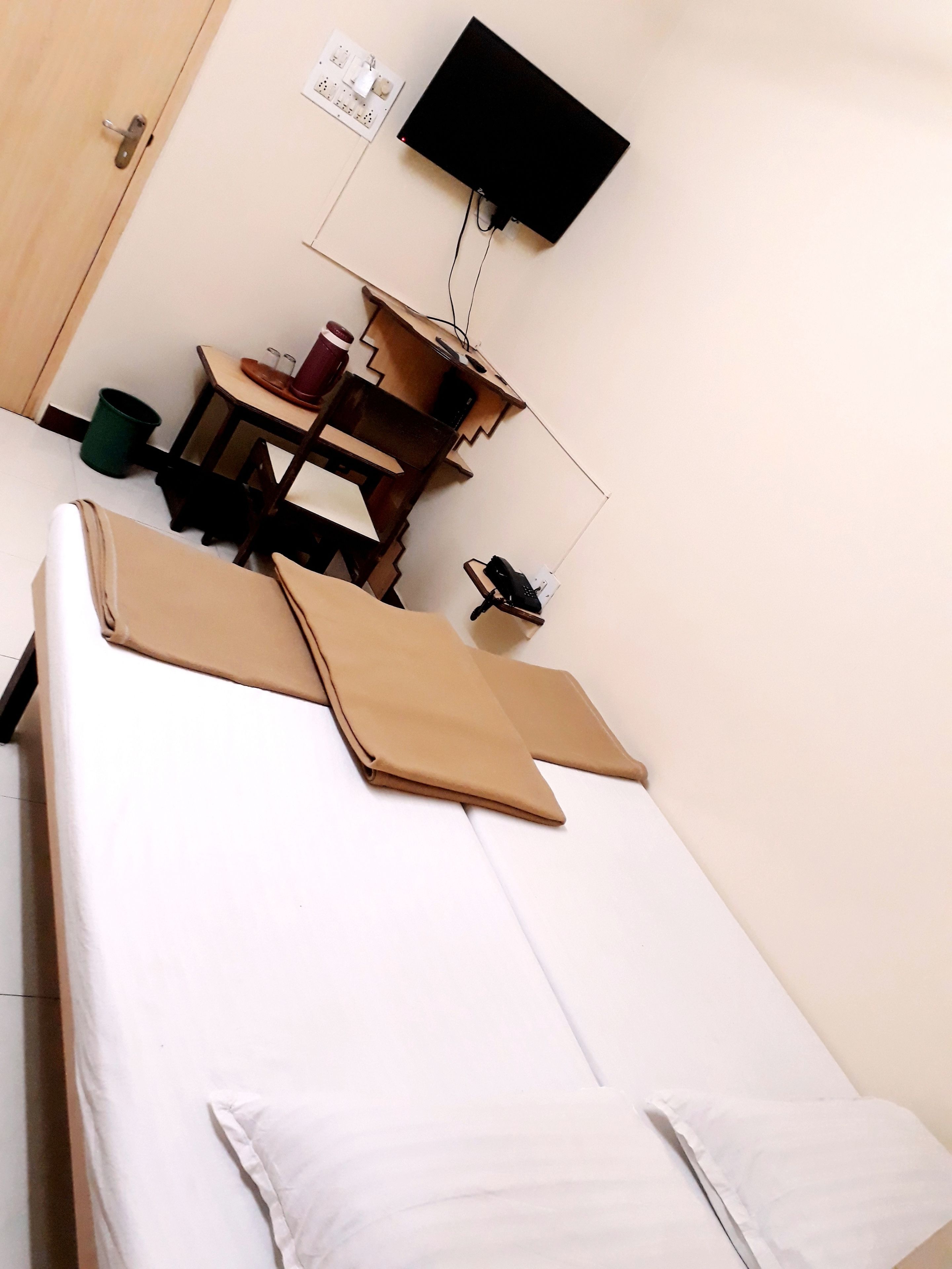 A single bed with a white mattress and pillows, a wooden desk with a chair, a small trash bin, and a wall-mounted TV.
