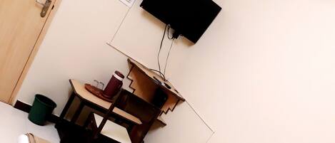 A single bed with a white mattress and pillows, a wooden desk with a chair, a small trash bin, and a wall-mounted TV.