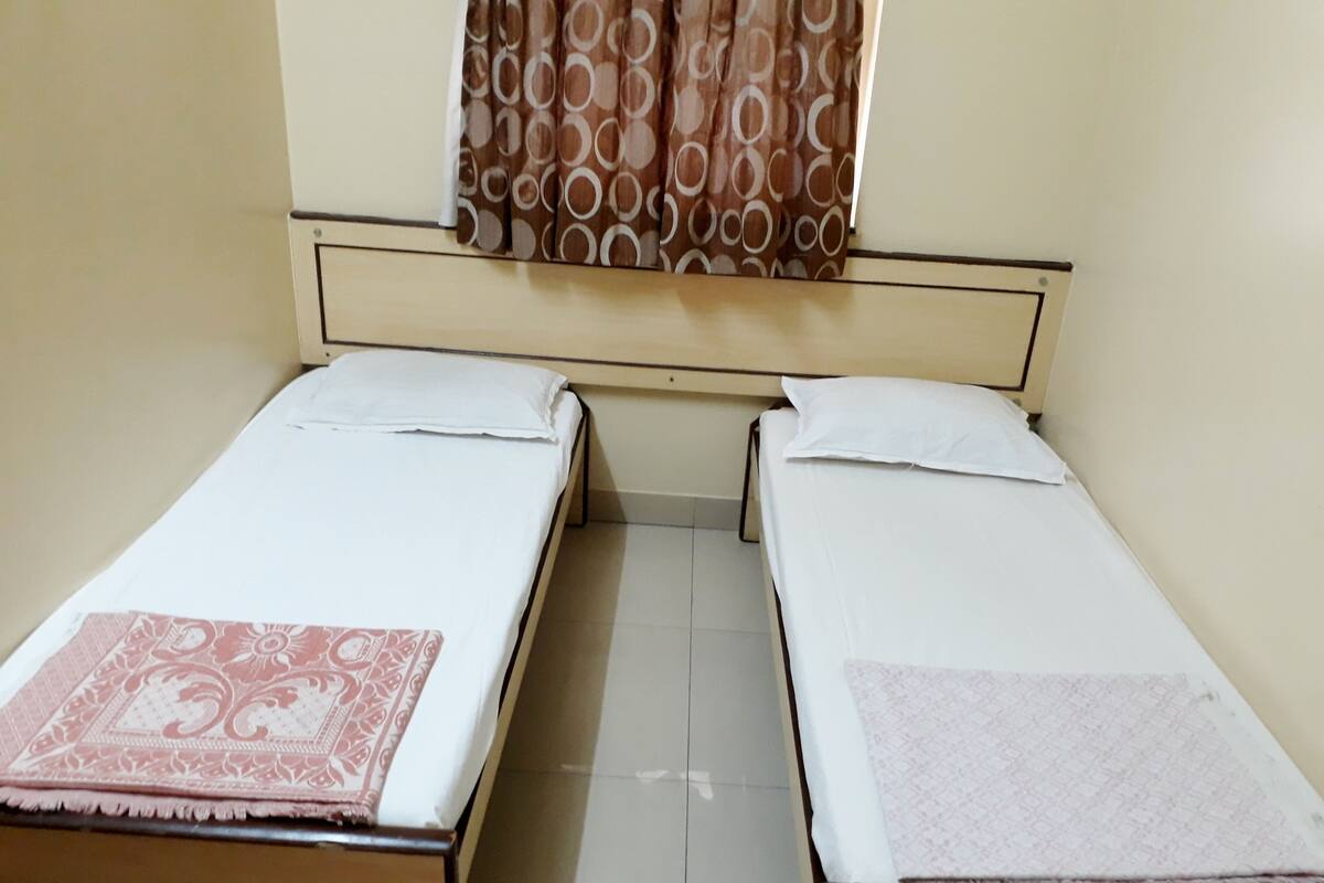 A small, single-room accommodation with two single beds, a window with patterned curtains, and a plain tiled floor.