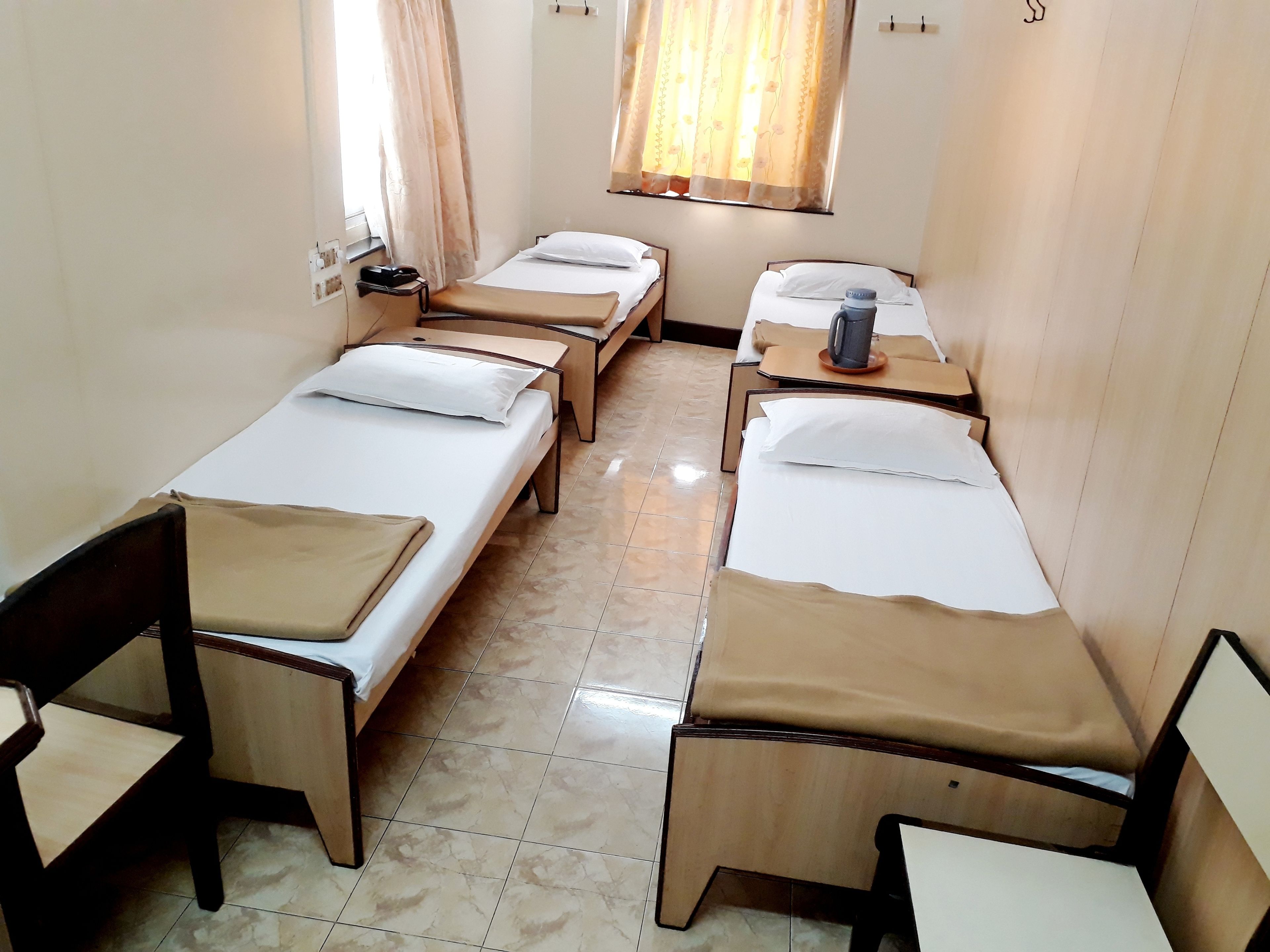 A hotel room with four single beds, each with a wooden frame and beige bedding. There are two bedside tables with lamps and a small table between the beds. The room has a tiled floor and a window with curtains.