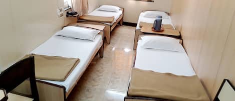 A hotel room with four single beds, each with a wooden frame and beige bedding. There are two bedside tables with lamps and a small table between the beds. The room has a tiled floor and a window with curtains.