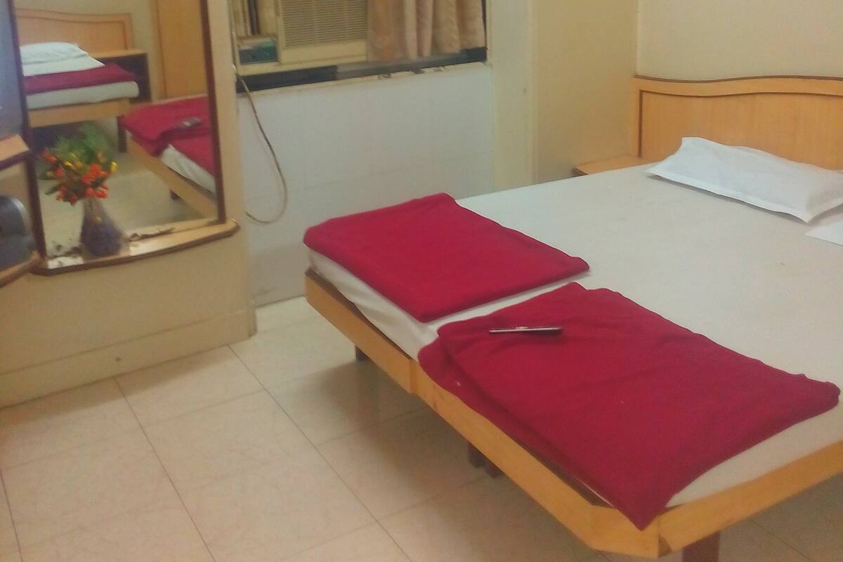 A small bedroom with a single bed, a bedside table with a mirror, a window with curtains, and a tiled floor.