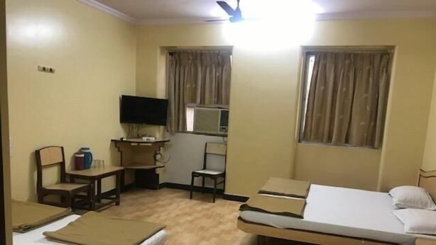 A hotel room with a bed, a desk with a chair, a television, and two windows with curtains. - Sangam Guest House (Mumbai)