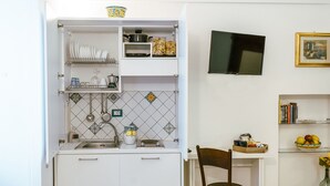 Una cucina compatta con mobili bianchi, zoccolo in piastrelle e un piccolo angolo ristoro con una sedia e un tavolo.