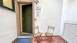 Piccola area d'ingresso chiusa, con una porta, due sedie pieghevoli, un tappetino d'entrata, una luce a parete e un cartello sopra la porta.