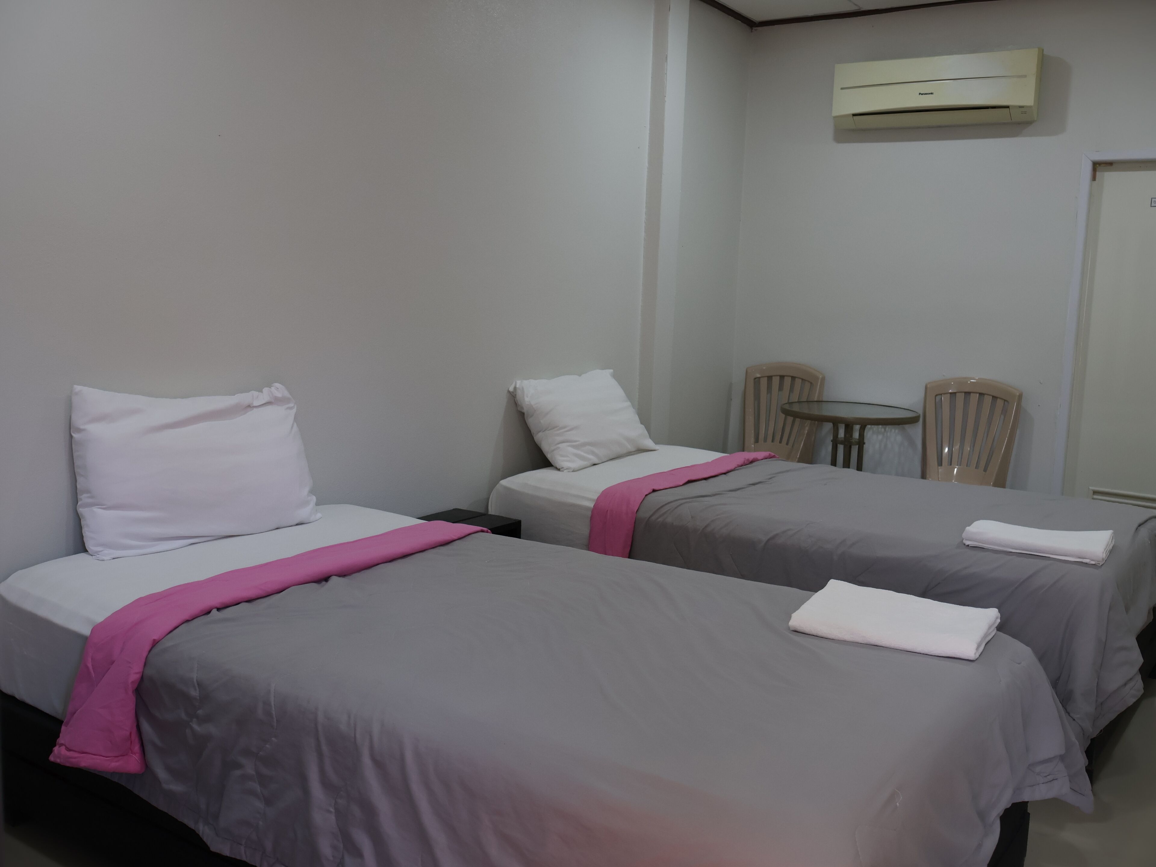 Hotelkamer met twee bedden, elk voorzien van een kussen en een roze deken. Er is een kleine tafel met twee stoelen, en een airconditioner aan de muur.