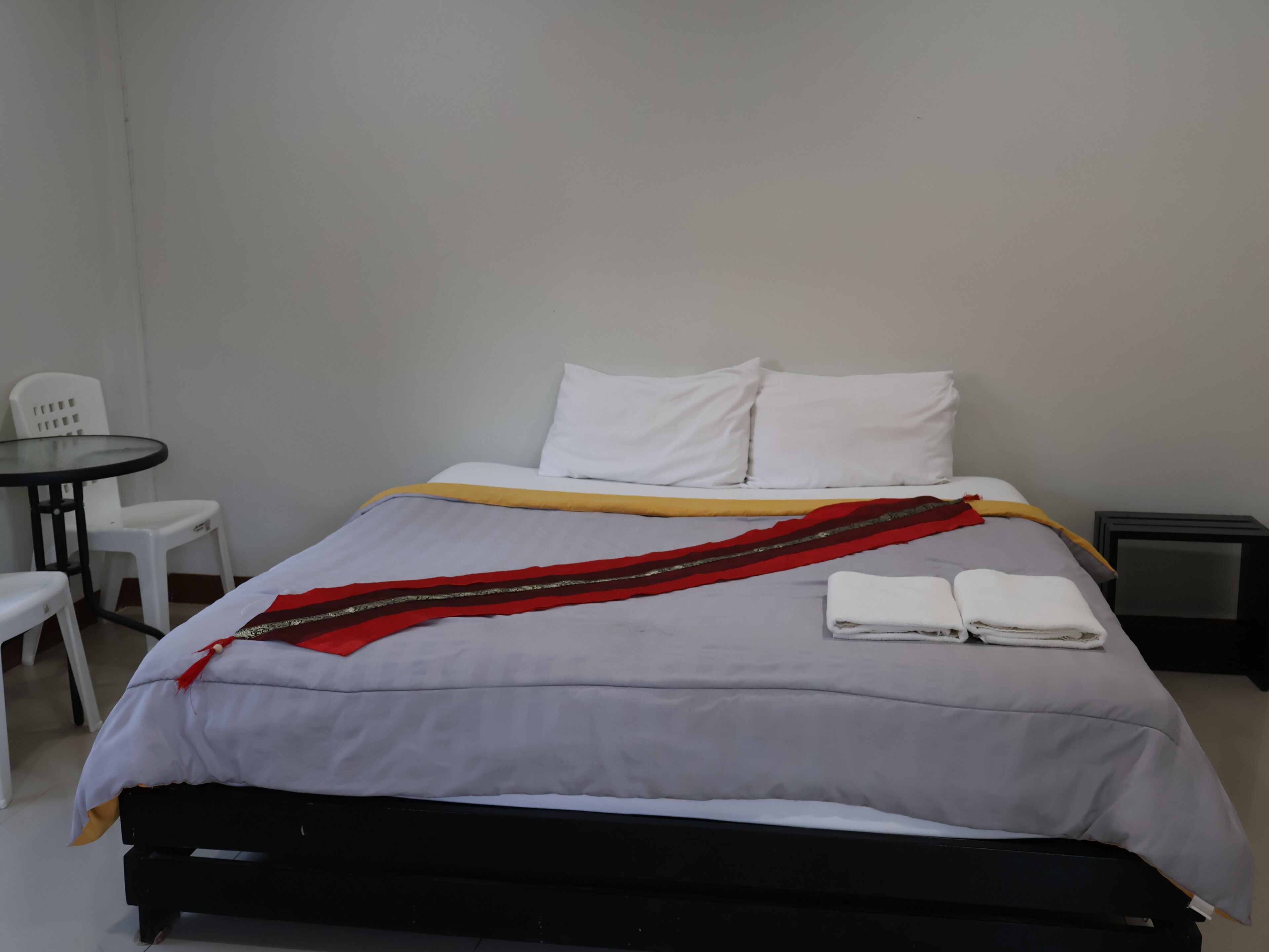 Camera d'albergo con un letto, cuscini bianchi, una coperta grigio e rossa, un tavolino con una sedia e un comodino.
