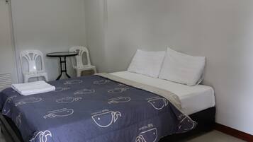 Une chambre d’hôtel avec un lit, deux chaises blanches, une petite table ronde et un climatiseur fixé au mur.