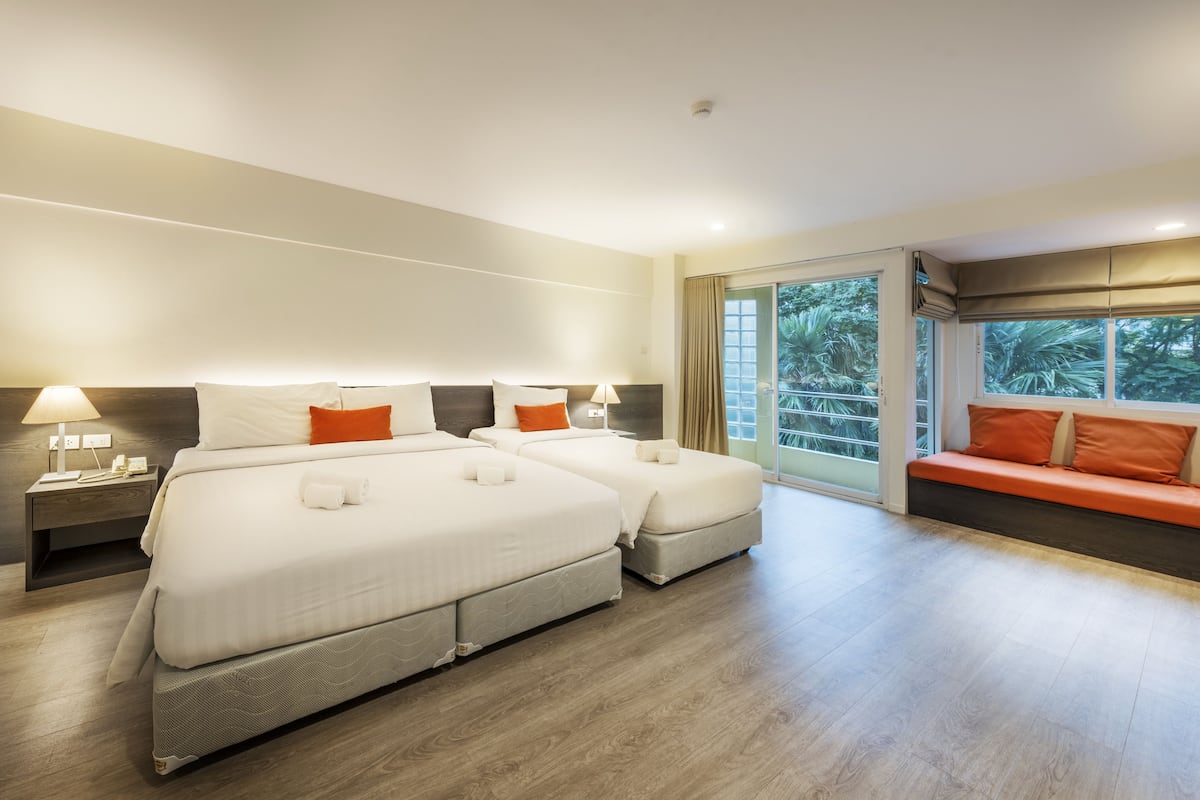 A hotel room with two beds, a sofa, a nightstand with a lamp, and a view of greenery through the window.