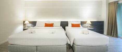 Habitación de hotel con dos camas, cada una con ropa de cama blanca y almohadas naranjas, un cabecero oscuro y una mesita de noche con una lámpara.