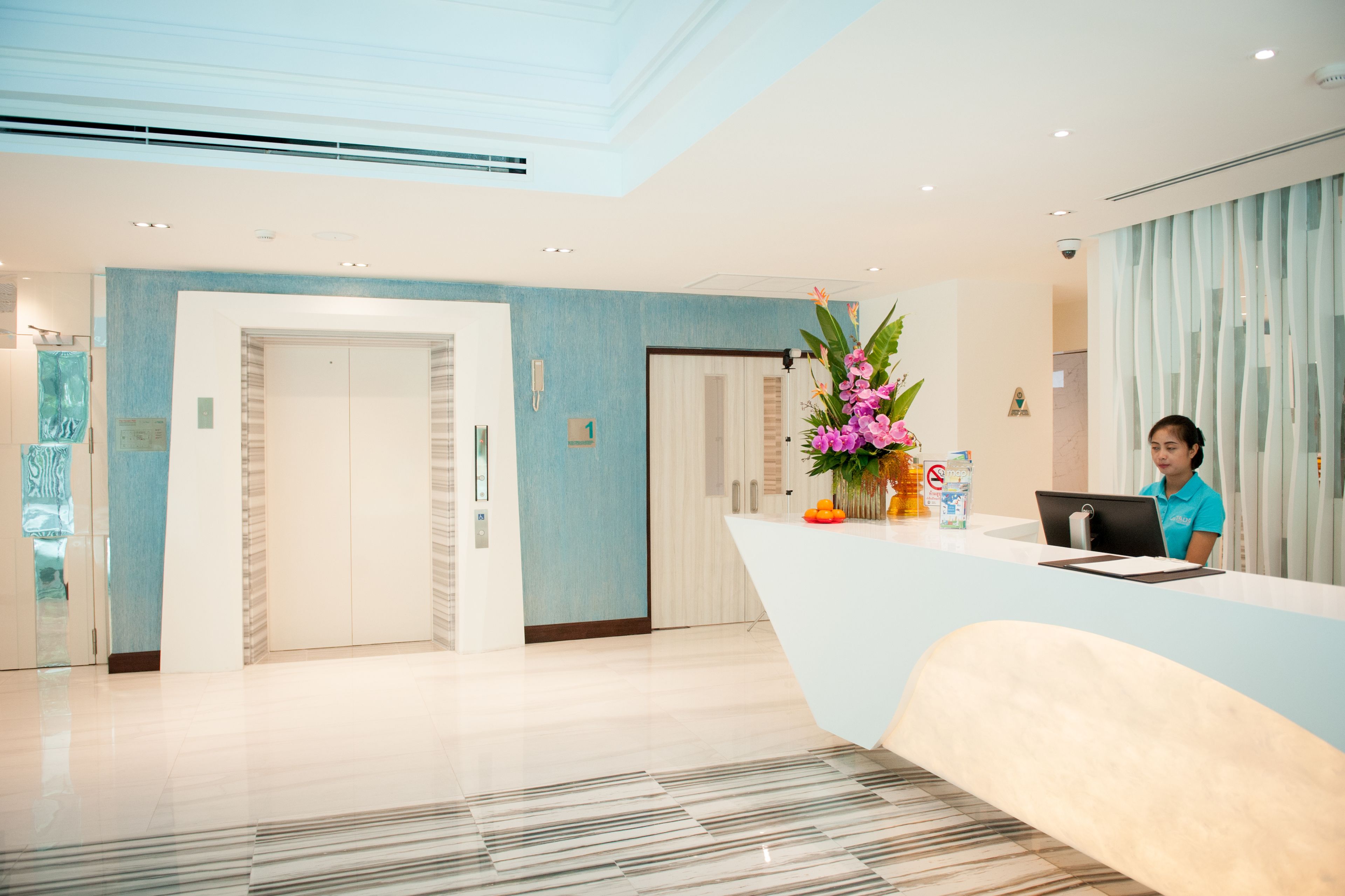 Uma área de recepção de hotel moderna, com um recepcionista no balcão, um grande arranjo de flores e um interior limpo e bem iluminado.