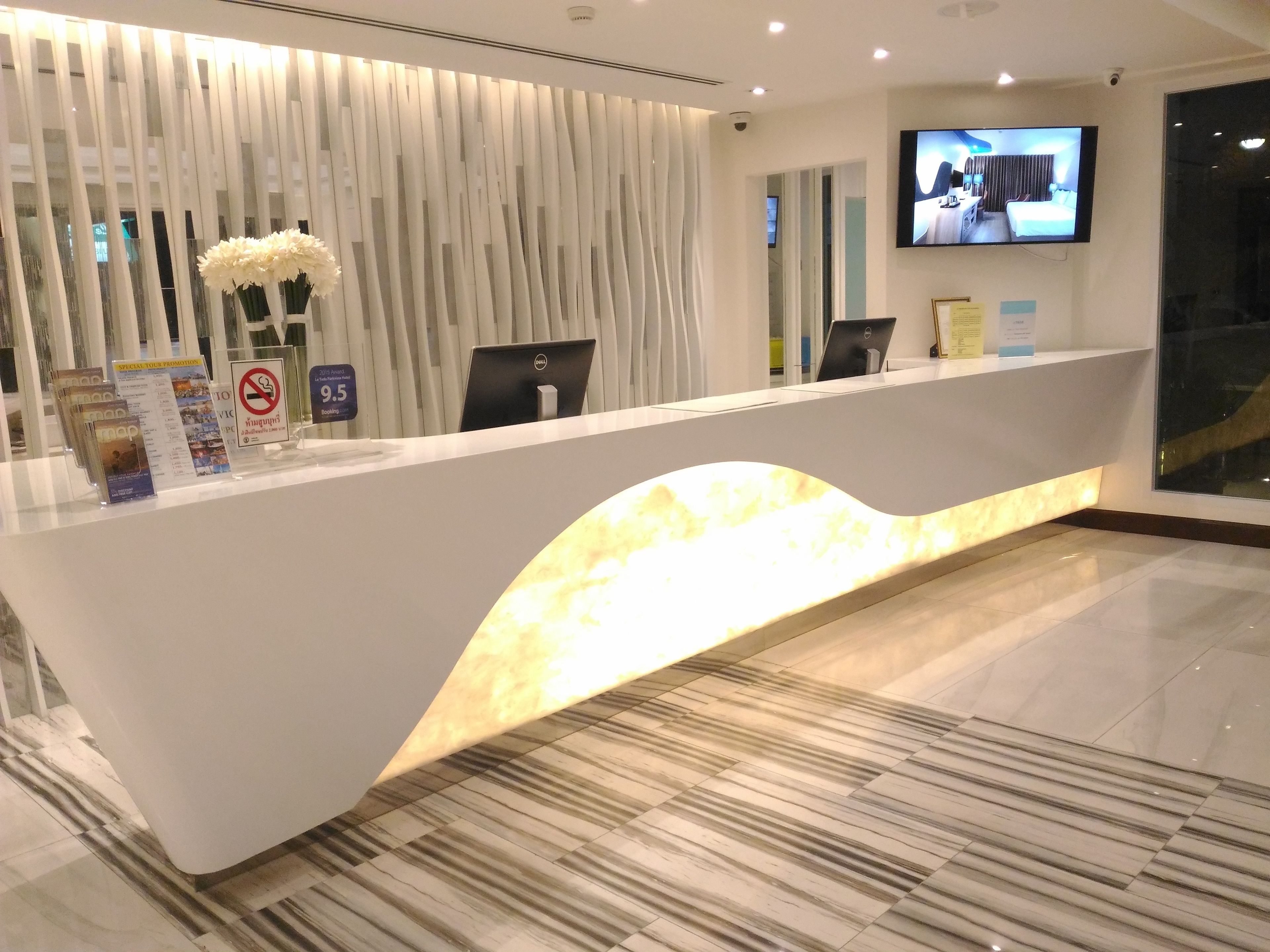 A hotel reception desk with a modern design, featuring a curved front and a lit-up base. There are two computer monitors, a potted plant, and informational brochures on the desk.