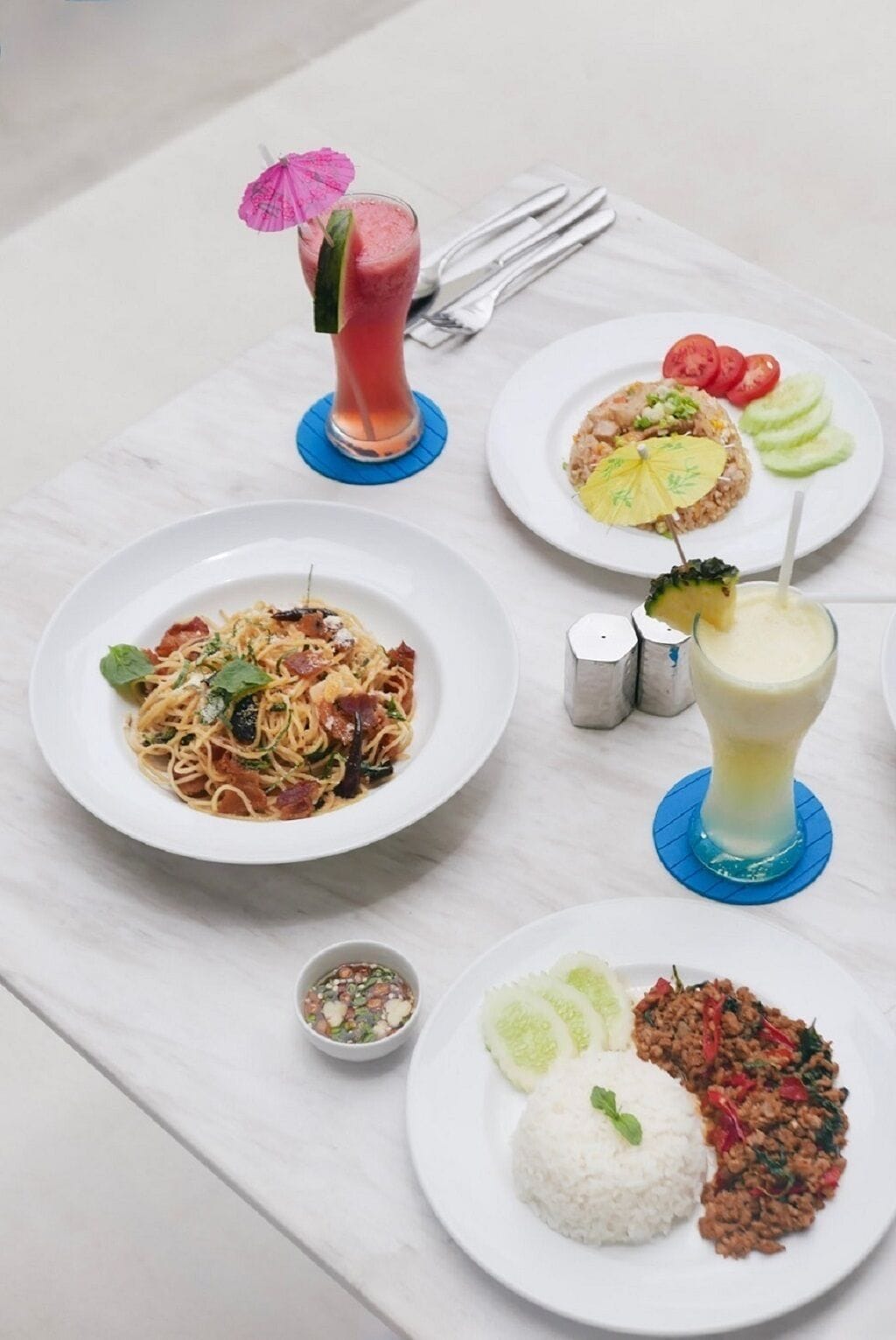 Une table où l’on trouve divers plats, notamment des pâtes, du riz accompagné de viande, et une boisson décorée d’une tranche d’ananas.