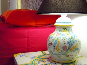 Una cama con una manta roja y un cojín, una lámpara sobre una mesita, y un florero colorido.