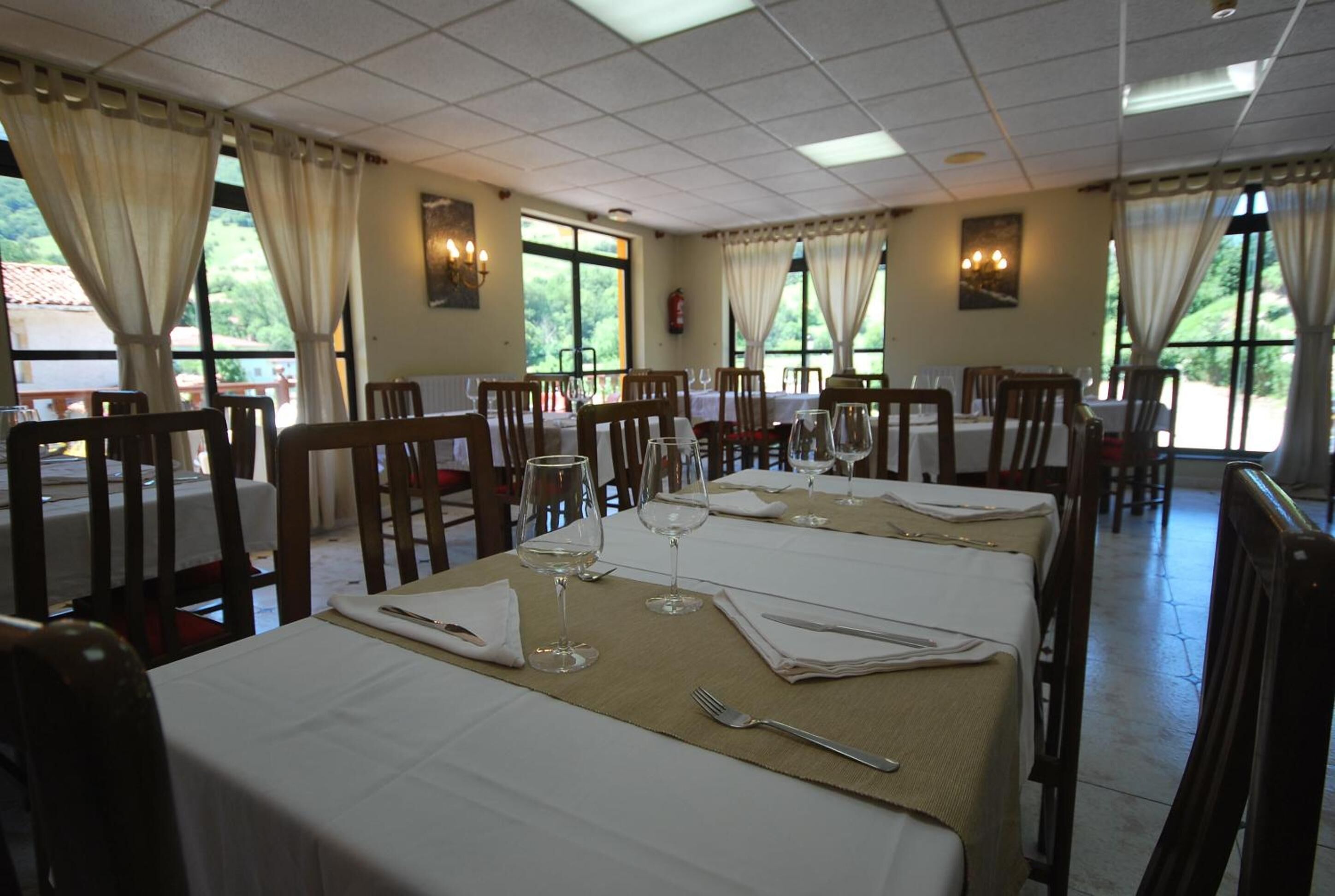 A dining area with tables set for four, each with a white tablecloth, wine glasses, and cutlery. The room has large windows with curtains, allowing natural light and views of greenery outside.