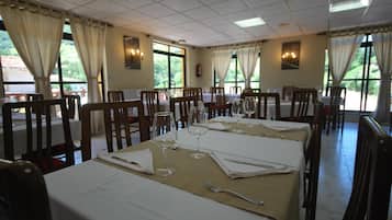 Un comedor con mesas dispuestas para cuatro, cada una con mantel blanco, copas de vino y cubiertos. La habitación tiene ventanas amplias con cortinas, que dejan entrar luz natural y ofrecen vistas a la vegetación exterior.