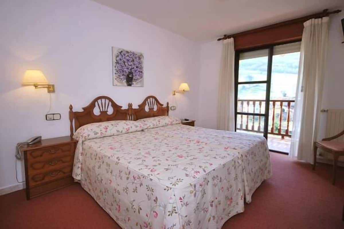 Une chambre à coucher avec un lit en bois, une couvre-lit à motifs floraux, deux lampes de chevet, une table de chevet et une fenêtre avec des rideaux.
