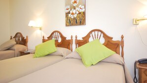 Ein Hotelzimmer mit zwei Betten, jeweils mit Holz-Kopfteilen und grünen Kissen. An der Wand hängt ein Blumenbild, links steht eine Lampe und rechts ein Telefon.
