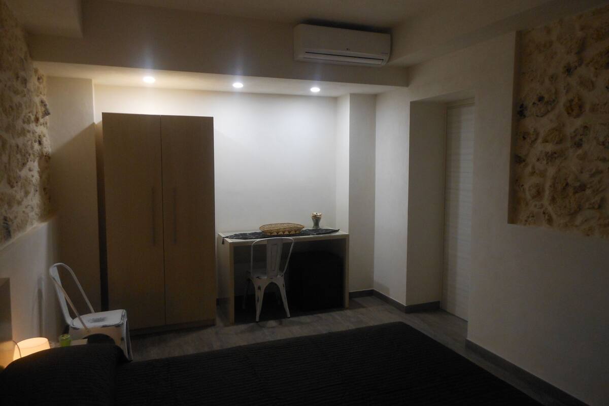 Una habitación pequeña y poco iluminada, con una cama, un escritorio, una silla y un equipo de aire acondicionado.