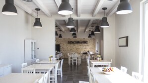 A spacious dining area with white tables and chairs, black pendant lights, and a stone wall in the background. - La Mela di Venere (Palazzolo Acreide)