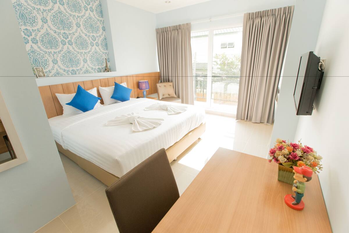 Ein Hotelzimmer mit einem großen Bett, einem hölzernen Kopfteil, einem kleinen Tisch mit Blumen in einer Vase und einem Balkon mit Vorhängen.