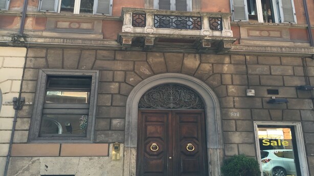 A building with a prominent entrance featuring a large wooden door with a decorative arch. There are multiple windows with shutters, and a small balcony with a railing. - QT Suites Roma (Rome)