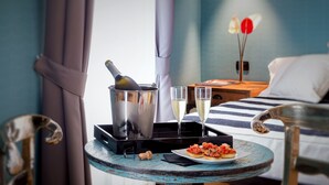A room with a bed, a bedside table with a bottle of champagne, two glasses of champagne, and a plate of food. - QT Suites Roma (Rome)