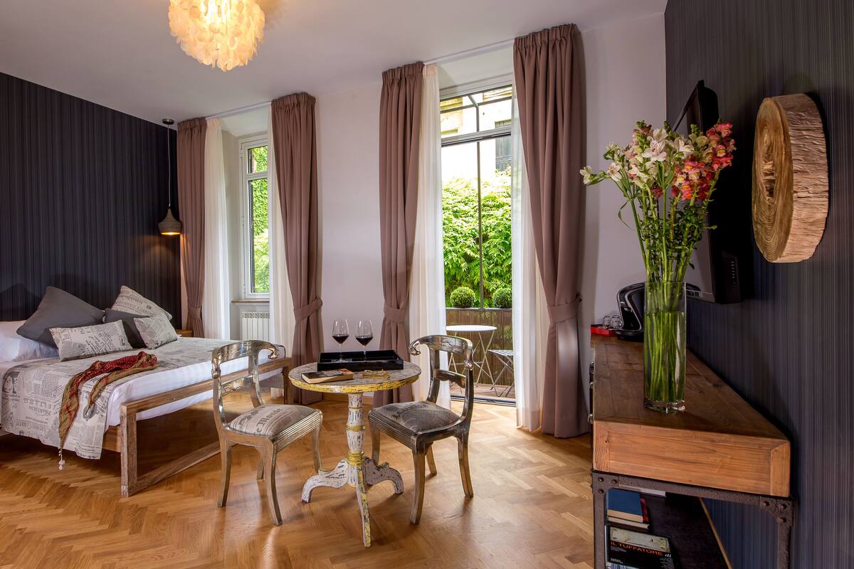 Een moderne slaapkamer met een groot bed, een klein rond tafeltje met stoelen, een boekenkast en uitzicht op groen.