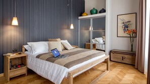 A modern bedroom with a wooden bed, a laptop on the bed, a bedside table with a book, and a nightstand with a lamp. - QT Suites Roma (Rome)