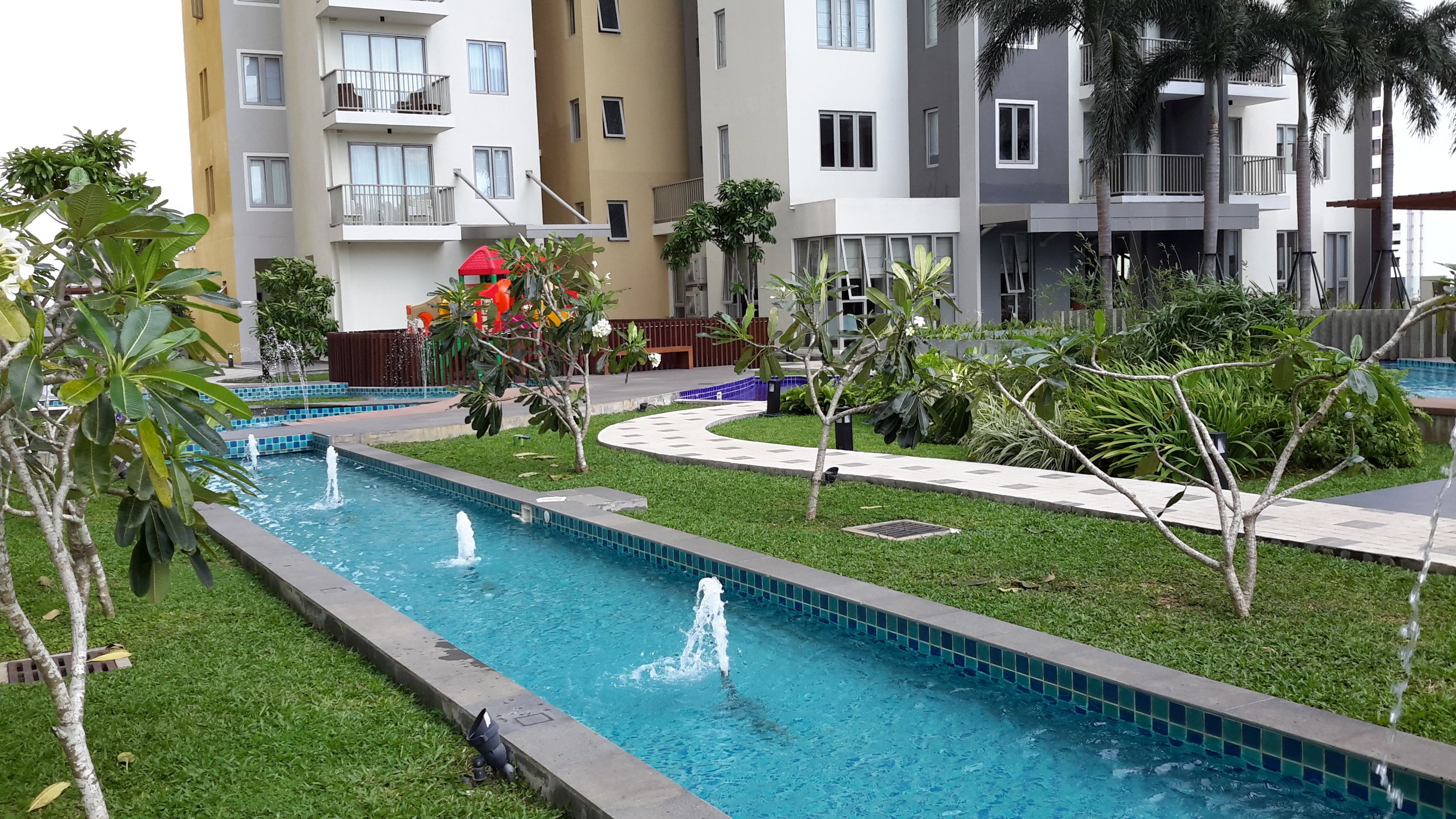 A swimming pool with multiple fountains, surrounded by a well-maintained lawn and various plants.