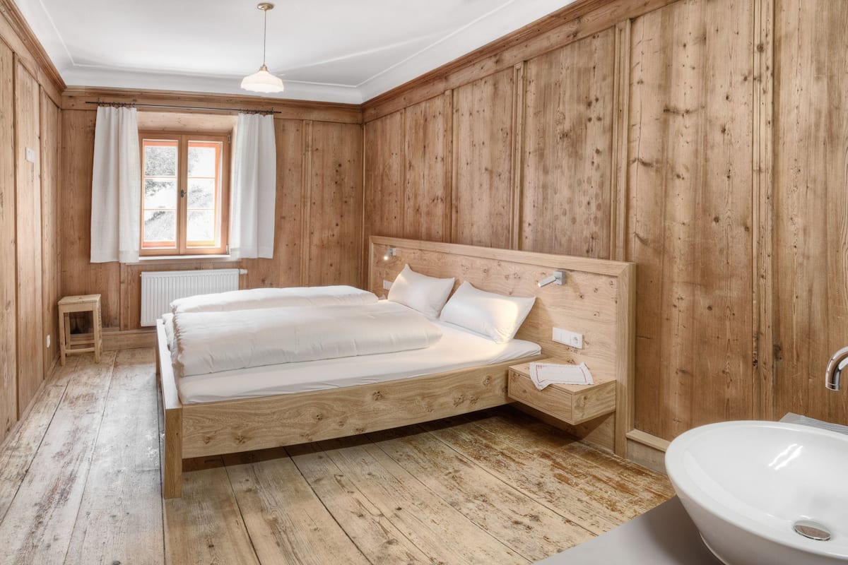 Une chambre à coucher aux murs lambrissés, avec un lit double, une petite table de chevet, une fenêtre avec des rideaux et un lavabo blanc.