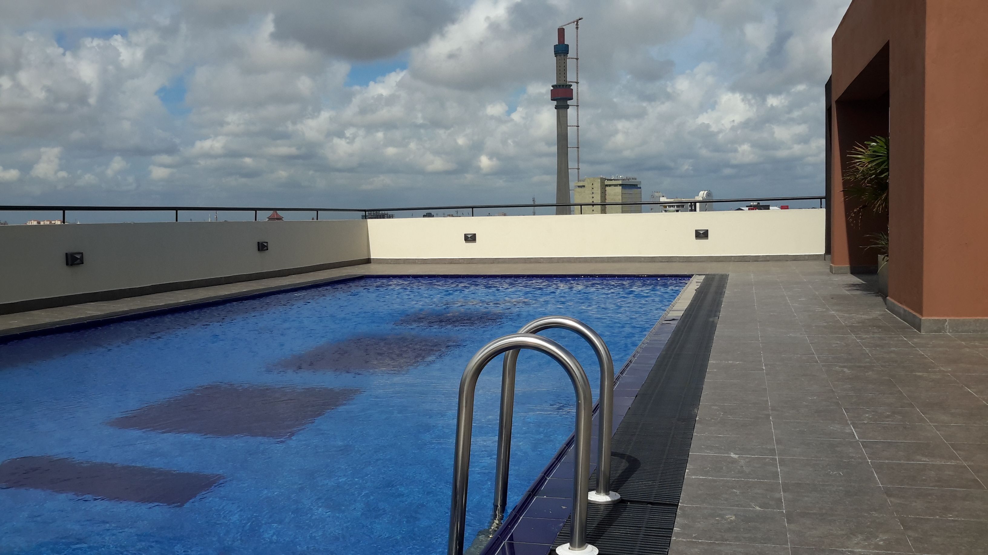 Una piscina en la azotea con barandillas metálicas, cielo despejado y edificios a lo lejos.