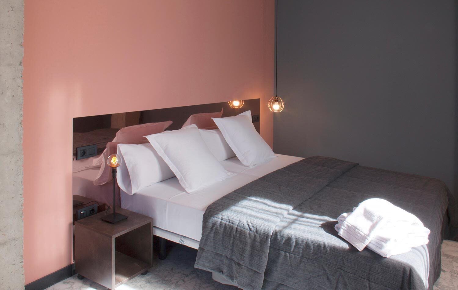 Un lit bien fait, avec une literie blanche et grise, une table de chevet avec une lampe et un luminaire fixé au mur.