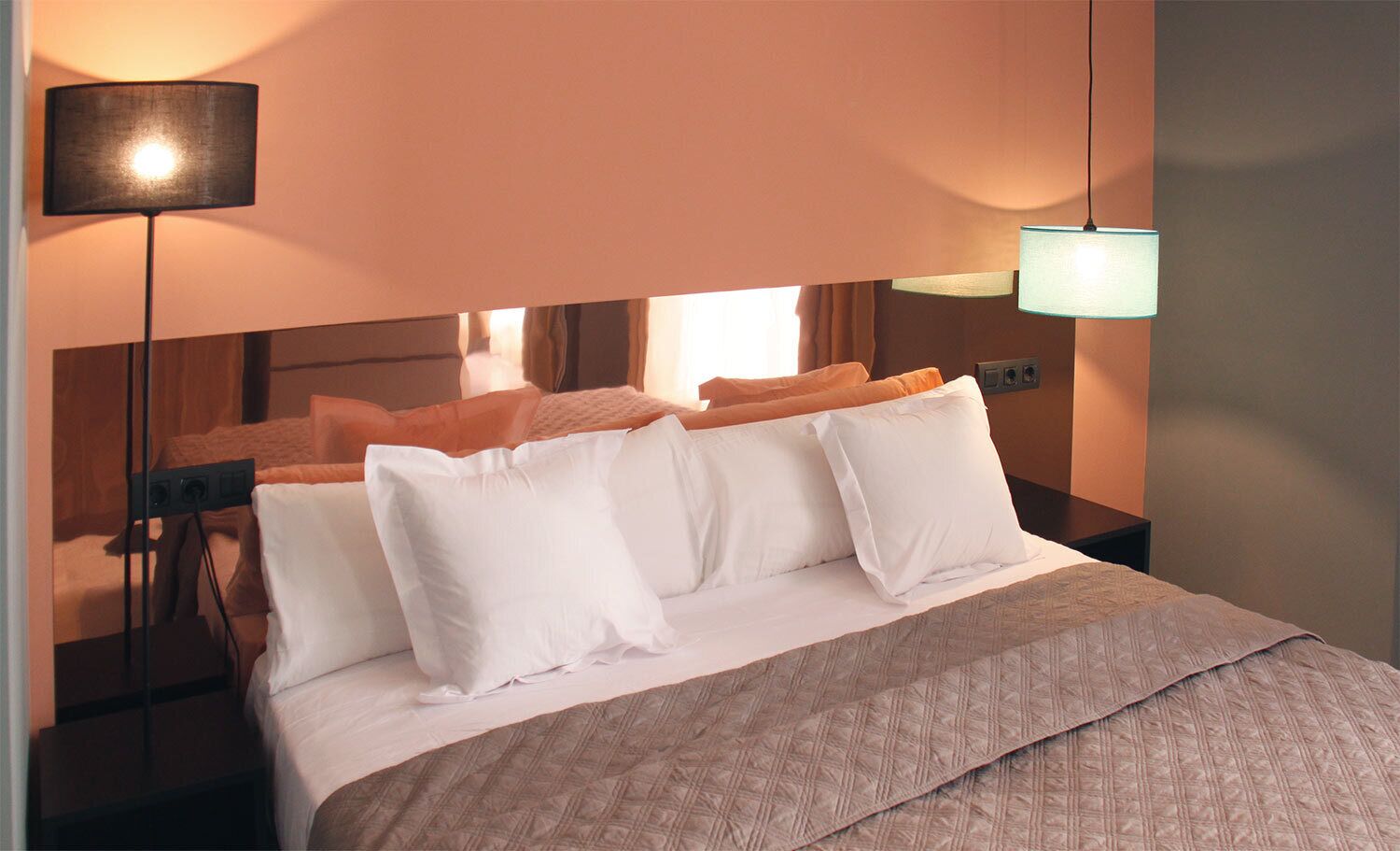 A hotel room with a bed, white pillows, a beige bedspread, a black floor lamp, and a wall-mounted light fixture.