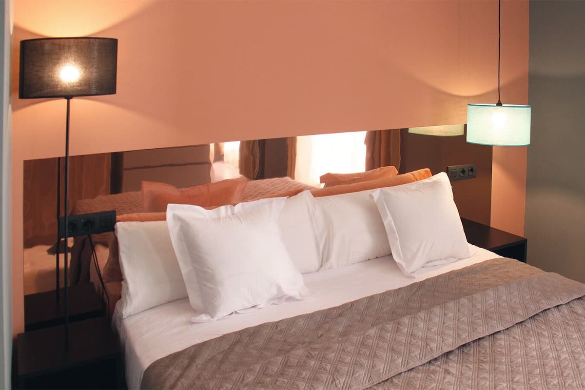 A hotel room with a bed, white pillows, a beige bedspread, a black floor lamp, and a wall-mounted light fixture.