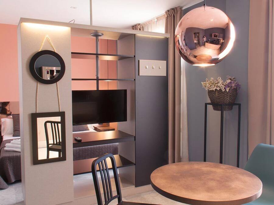 Quarto de hotel moderno com televisão, mesa redonda, cadeira, espelho e planta decorativa.