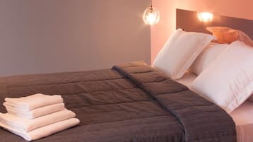 Una cama bien hecha con una colcha marrón, almohadas blancas y toallas dobladas a un lado. Dos lámparas de pared están sobre la cama.