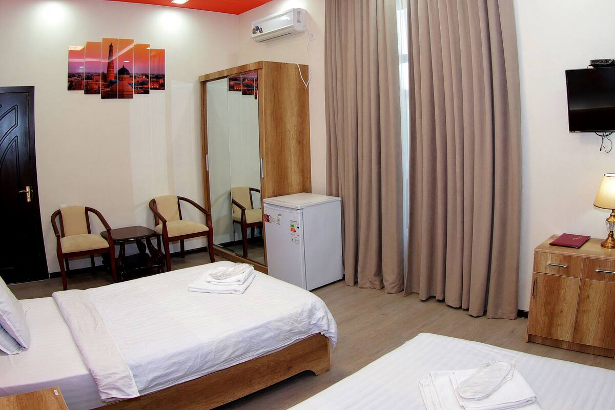 A hotel room with two beds, a desk, a chair, a minibar, a TV, and a large window with curtains.