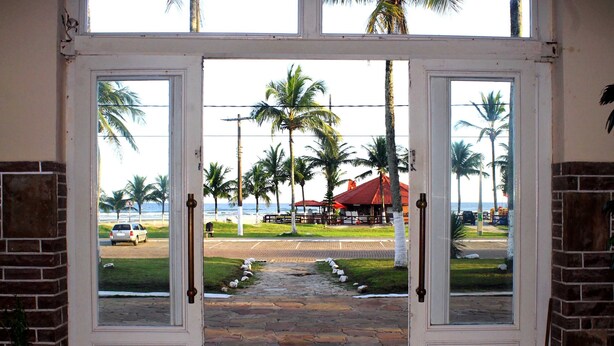 A doorway leading to a beachside area with palm trees and a red-roofed building. - Hotel H Itanhaém (Itanhaém)