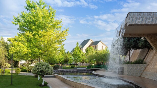 Ein angelegter Garten mit einem Brunnen, einem Baum und einem Gebäude im Hintergrund.