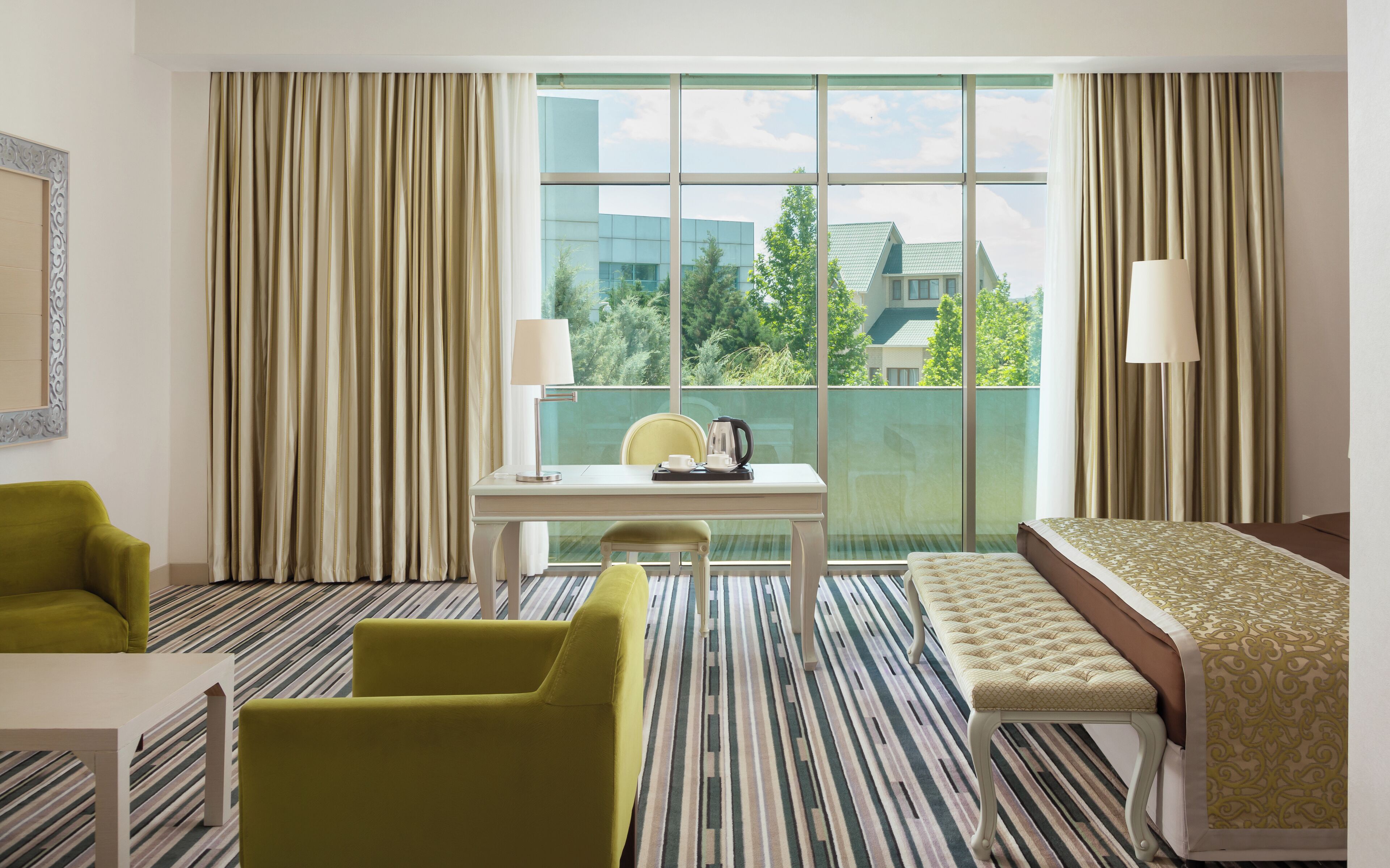 A hotel room with a large window, a bed, a desk with a chair, a small table, and a green armchair.