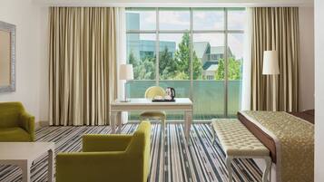 A hotel room with a large window, a bed, a desk with a chair, a small table, and a green armchair.