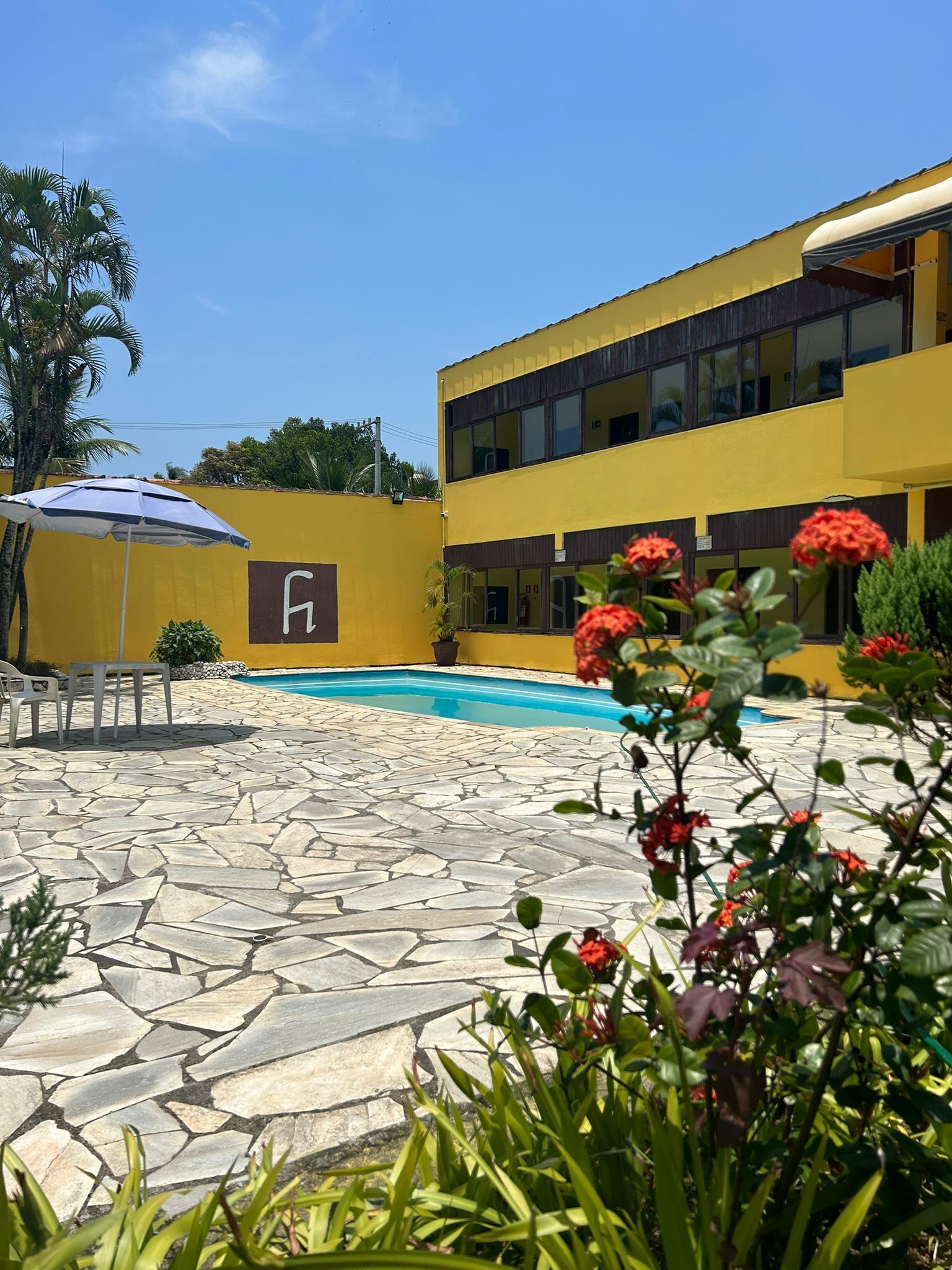 Zona de piscina con baldosas de piedra, un edificio amarillo con letrero 'H' y flores rojas en primer plano.